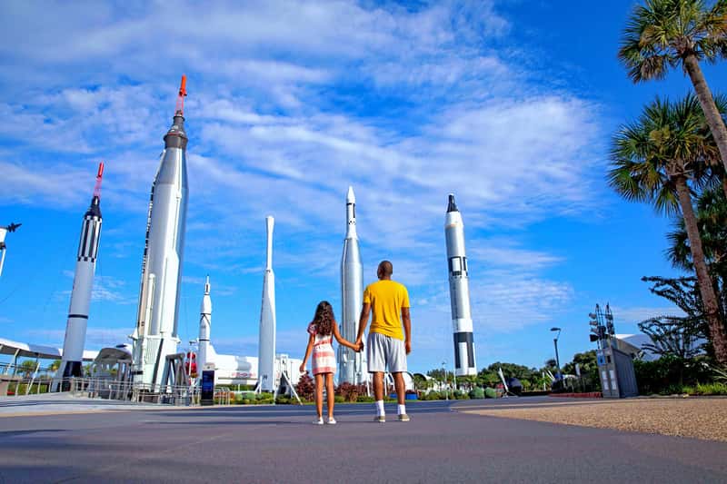 A father and daughter hold hands looking up at the Kennedy Space Center